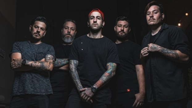 Eine Gruppe von Männern, die in einem schwach beleuchteten Raum stehen, von rechts beleuchtet, während sie offenbar ein Interview für Avenged Sevenfold geben.