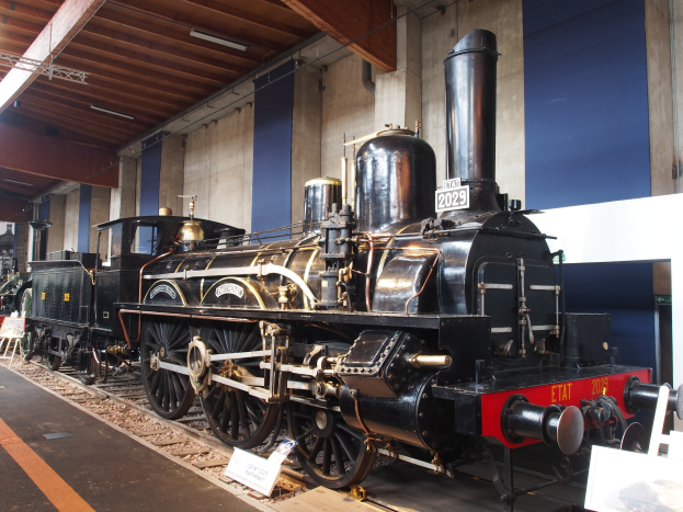 Eine nostalgische Dampflokomotive in einem Museum ausgestellt, umgeben von Eisenbahnschienen, Informationsschildern und anderen Ausstellungen, mit einer beleuchteten Decke und Wand im Hintergrund.