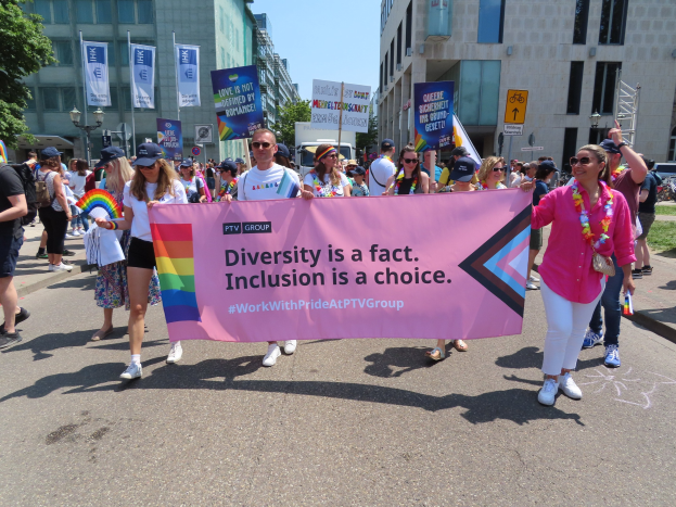 Eine bunte Gruppe von Menschen marschiert auf der Stra├če, h├Ąlt ein Banner mit der Aufschrift "Diversit├Ąt ist eine Tatsache, Inklusion ist eine Wahl", einige tragen Mützen und Schutzbrillen, andere halten zus├Ątzliche Banner, vor einem Hintergrund von Geb├Ąuden, B├Ąumen, Laternenmasten und einem klaren blauen Himmel.