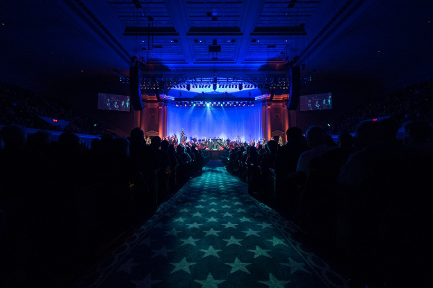 Ein großer Auditoriumsaal mit Sitzenden, eine Bühne mit Musikern und eine Decke mit Lichtern und Fernsehgeräten, was auf einen Konzertsaal für die Nationale Konferenz 2019 hindeutet.