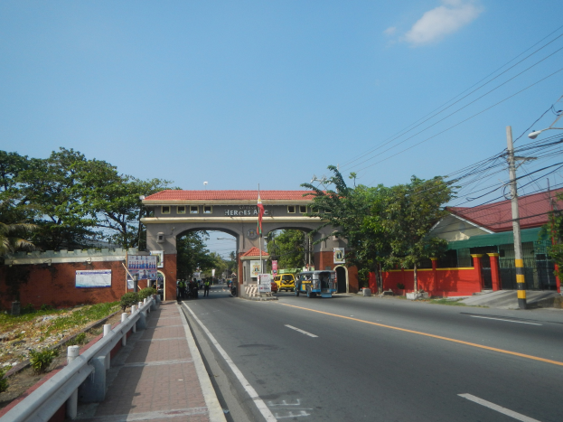 Eine Straße mit fahrenden Autos, ein Fußweg mit Geländern auf der linken Seite, Bäume und Gebäude auf beiden Seiten der Straße, ein Tor mit einer Namensschilder, Strommasten mit Drähten und ein bewölkter Himmel im Hintergrund.
