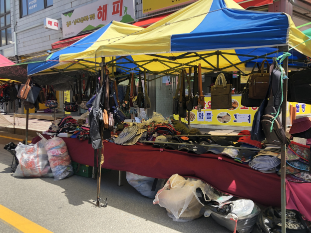 Ein Straßenmarkt mit Zelten, die Taschen, Hüte und andere Gegenstände präsentieren, vor einem Hintergrund aus Gebäuden mit Fenstern und Texttafeln.