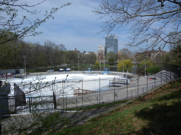 Eissporthalle umgeben von einem Metallzaun, Gras, Bäumen, Fahrzeugen, Straßenlaternen, einem Gewässer, Gebäuden und einem bewölkten Himmel.