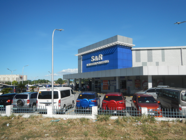 Ein Autohaus mit parkenden Fahrzeugen vorne, umgeben von einem Metallzaun, Gras, Pflanzen, Bäumen, Straßenlaternen und einem bewölkten Himmel.