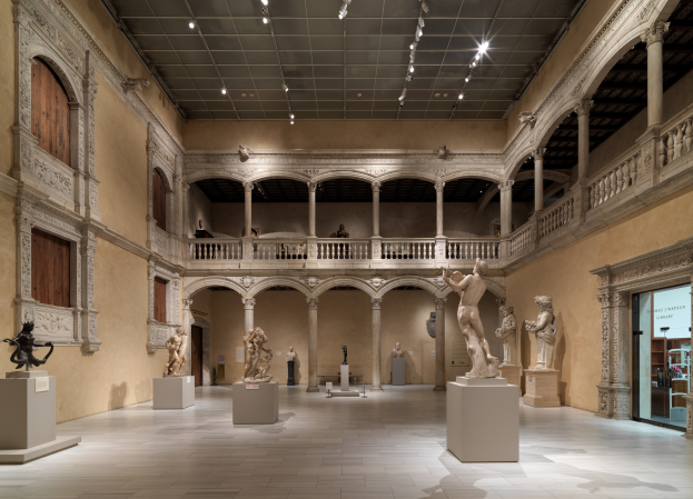 Ein geräumiger Museumsgalerie mit zahlreichen Statuen und Skulpturen auf Sockeln, mit architektonischen Elementen wie Säulen, Geländern und einer beleuchteten Decke sowie einigen Besuchern, die die Kunstwerke betrachten.