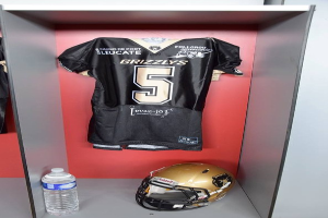 Ein Fussballtrikot und Helm hängen an einer Spindtür, mit einer Wasserflasche auf dem Regal darunter.