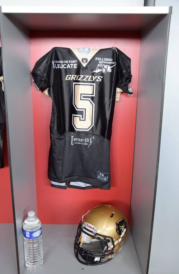 Ein Fussballtrikot und Helm hängen an einer Spindtür, mit einer Wasserflasche auf dem Regal darunter.