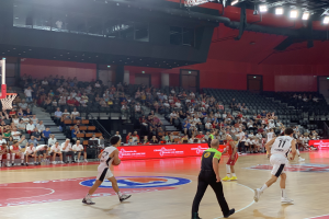 Eine Gruppe von Menschen, die Basketball in einer Turnhalle spielt, mit einem Basketballkorb links, Zuschauern im Hintergrund und beleuchteter Arena.