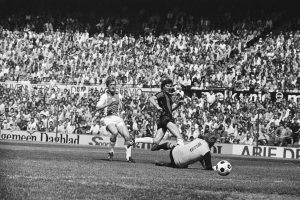Schwarzes Foto von Männern, die auf einem Feld Fußball spielen, mit Zuschauern im Hintergrund und einer Bundesliga 1978-79 Ajax 1-1 Hannover 96-Banner.