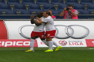 Zwei Fußballspieler feiern ein Tor auf einem Feld, umgeben von einer Gruppe von Menschen, mit einer Person, die eine Kamera hält, einem Banner mit Text, Stühlen und einer Wand im Hintergrund.