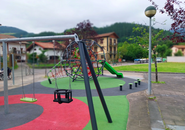 Spielplatz mit Schaukeln, einer grünen Rutsche, Pfählen, Laternen, Bänken, Gras, Bäumen, Gebäuden, Fahrzeugen, Mülltonnen, Hügeln und einem klaren blauen Himmel.