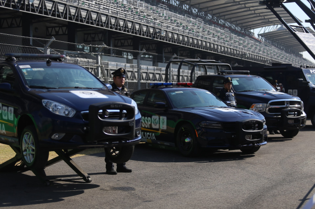 Polizeiautos vor einer Rennstrecke mit uniformierten Personen, Stadion und Zaun im Hintergrund.