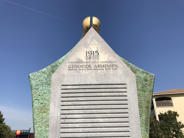 Ein hoher Denkmal mit einer Uhr darauf, beschriftet mit Text, steht in einem Park umgeben von Bäumen und Gebäuden unter einem klaren Himmel und erinnert an den 24. Jahrestag des Völkermords in Armenien.