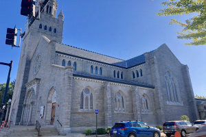 Große steinerne Kirche mit einem Glockenturm, identifiziert als St. Mary's Episcopal Church, umgeben von Gebäuden, Fahrzeugen, Fußgängern und Grünflächen unter einem bewölkten Himmel.