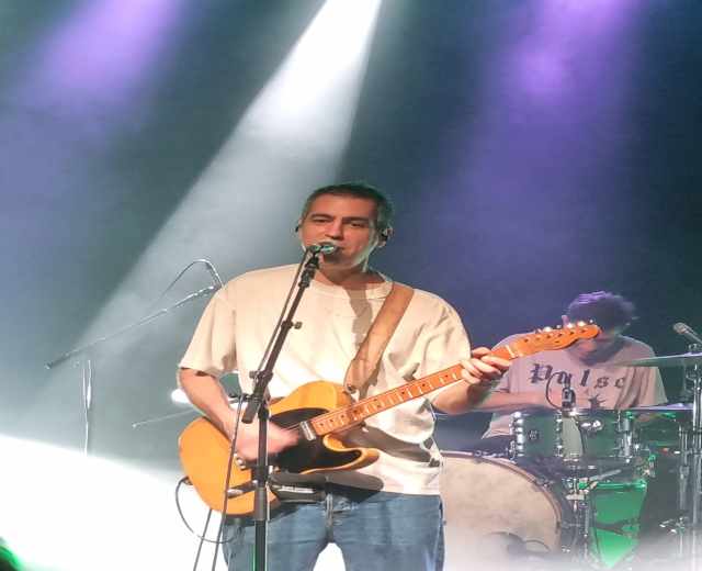 Ein Mann spielt eine E-Gitarre auf der Bühne und singt in ein Mikrofon, während ein Schlagzeuger hinter ihm sitzt.