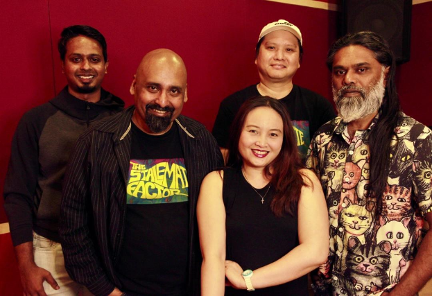 Eine Gruppe von Menschen, die in einem Aufnahmestudio zusammenstehen, lächeln und für ein Foto vor einer roten Wand posieren.