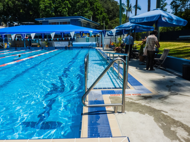 Großes Freibad mit schwimmenden Menschen, umgeben von Geländern, Sonnenschirmen, Stühlen, Tischen, Pflanzen, Gras, Bäumen und einem Gebäude, unter einem klaren blauen Himmel.