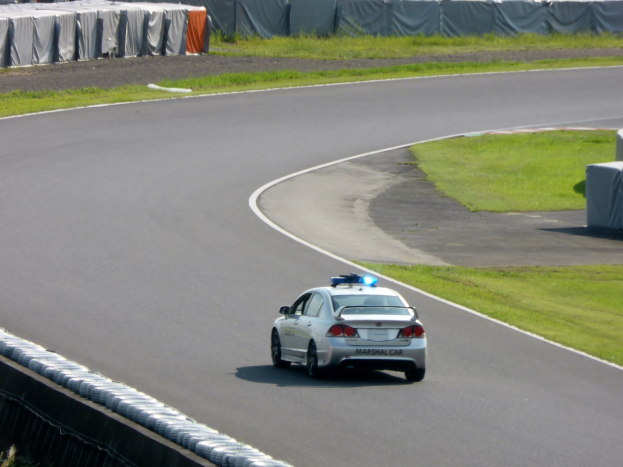 Polizeiauto fährt auf einer Rennstrecke mit Gras auf beiden Seiten, im Hintergrund sind Menschen und Zelte zu sehen.