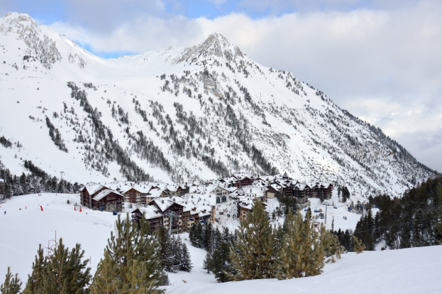 Eine Winterlandschaft in einem Skigebiet mit schneebedeckten Bergen, Bäumen, die von Schnee bedeckt sind, einer Hütte zwischen den Bäumen und einem bewölkten Himmel.