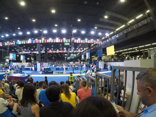 Eine große Menschenmenge sitzt und steht in einer Indoor-Sportarena mit Bildschirmen, Fahnen und Deckenleuchten im Hintergrund, getrennt von der Bühne durch einen Metallzaun.