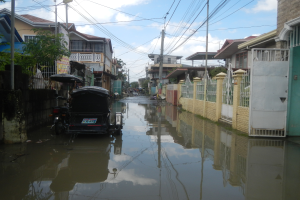Eine überflutete Straße mit einem Motorrad in der Mitte, Gebäude, Bäume und Strommasten mit Drähten auf beiden Seiten sowie einige Menschen und Fahrzeuge im Hintergrund.