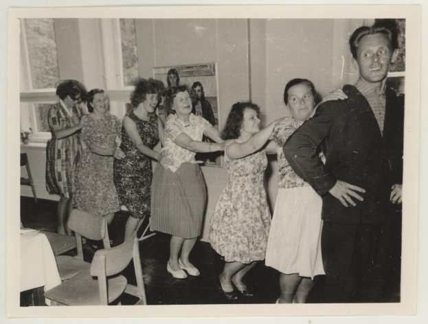 Eine Gruppe von Menschen, die in einem Raum mit Stühlen und einem mit einer wei├čen Decke bedeckten Tisch tanzen. An der Wand h├Ąngt ein Foto und im Hintergrund sind Fenster zu sehen.