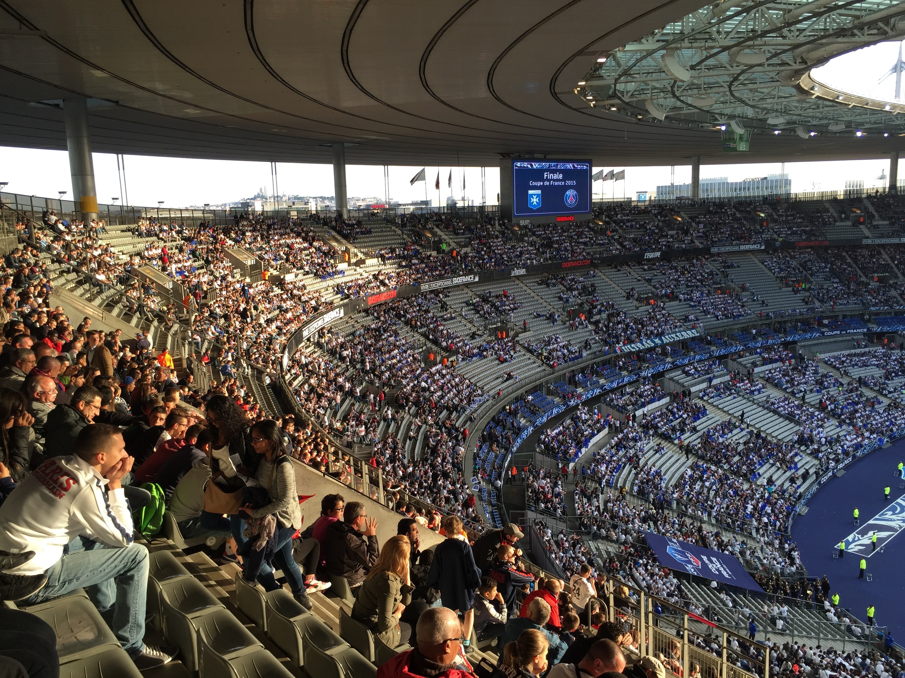 Große Menschenmenge in einem Stadion bei einem Fußballspiel mit einer Bühne rechts, Fahnen, Stangen, einem Bildschirm und dem Allianz Stadion in München, Deutschland im Hintergrund.