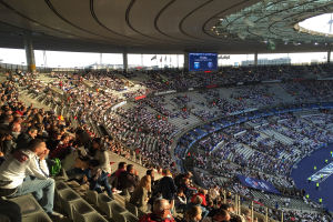 Große Menschenmenge in einem Stadion bei einem Fußballspiel mit einer Bühne rechts, Fahnen, Stangen, einem Bildschirm und dem Allianz Stadion in München, Deutschland im Hintergrund.