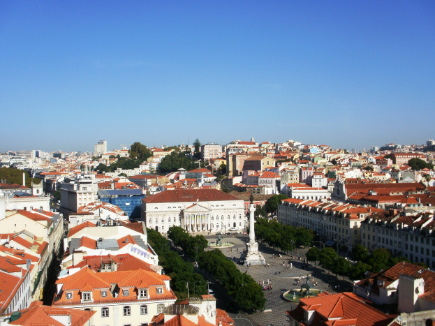 Blick auf Lissabon von einem Hügel mit Gebäuden, Bäumen, einer Statue auf einem Sockel, einigen Menschen auf der Straße und dem Himmel im Hintergrund.
