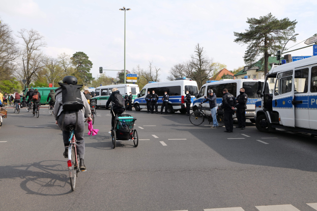 Eine Gruppe von Menschen auf Fahrrädern auf einer Straße mit Polizeiwagen und anderen Fahrzeugen, einige tragen Helme und haben Taschen oder schieben Kinderwagen, vor einer Kulisse aus Bäumen, Gebäuden und Verkehrsinfrastruktur.