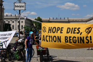 Eine Gruppe von Menschen steht vor einem Gebäude und hält ein gelbes Banner in der Hand, auf dem "Hope Dies, Action Begins" steht. In der Nähe parken mehrere Fahrräder und es gibt einen Pfahl mit einer daran befestigten Tafel, vor einem klaren blauen Himmel mit Bäumen im Hintergrund.
