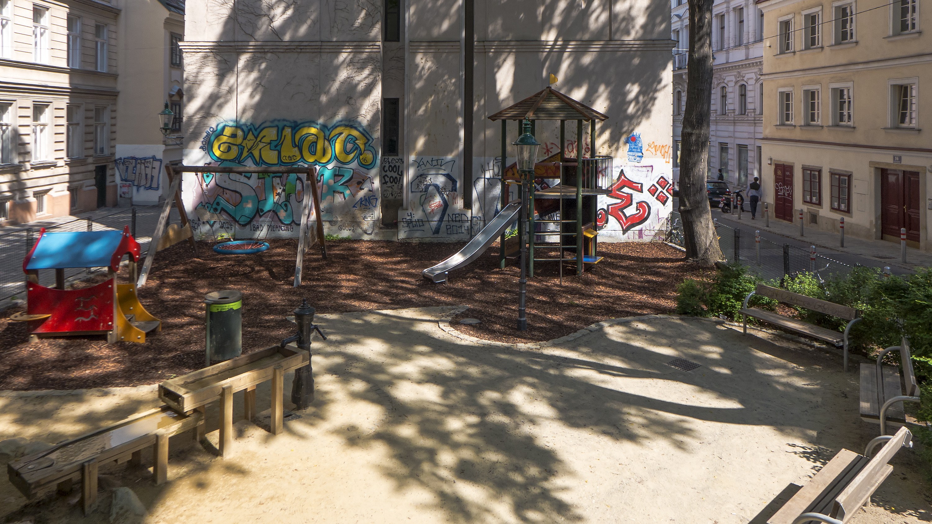 Spielplatz mit Graffiti in einer städtischen Umgebung, umgeben von Gebäuden, Bäumen, Bänken und einem Mülleimer, mit Menschen und Fahrzeugen auf der Straße und einem sichtbaren Himmel im Hintergrund.