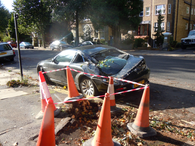Ein besch├Ądigt aussehendes Auto am Stra├čenrand mit orangefarbenen Verkehrskegeln drumherum, vor dem Hintergrund von B├Ąumen, Geb├Ąuden und einem klaren blauen Himmel.