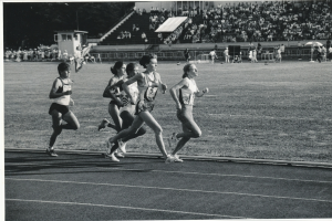 Eine Gruppe von Frauen, die auf einer Laufbahn in einem Stadion laufen, mit einer Zuschauermenge im Hintergrund, alles in Schwarz-Weiß dargestellt.