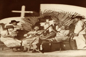 Ein Schwarz-Weiß-Foto einer Gruppe von Menschen, einige tragen Hüte und halten Musikinstrumente, die vor einer Kirche mit einem Kreuz an der Wand sitzen, betitelt mit "Mariachi Band, San Antonio, Texas".