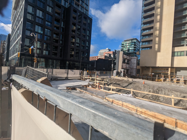 Baustelle in Toronto, Ontario mit einem großen Bauwerk im Bau, umgeben von einem Zaun, Pfählen, Verkehrszeichen und Schildern unter einem bewölkten Himmel.