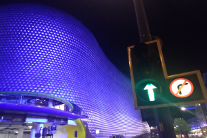 Das Selfridges Einkaufszentrum in Birmingham, England, ein Gebäude mit Säulen und Lichtern, umgeben von Bäumen, Laternenmasten und Fahrzeugen auf der Straße, mit einer Ampel vorne und dem Himmel im Hintergrund.