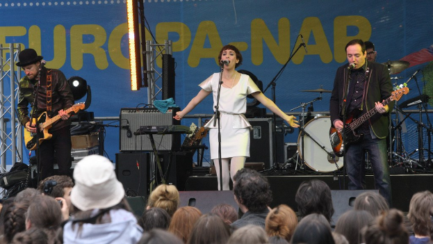 Vier Musiker treten auf der Bühne auf: eine Frau in einem weißen Kleid singt in der Mitte, flankiert von zwei Gitarristen, mit einer Menge unten und Lautsprechern an einer Wand im Hintergrund.