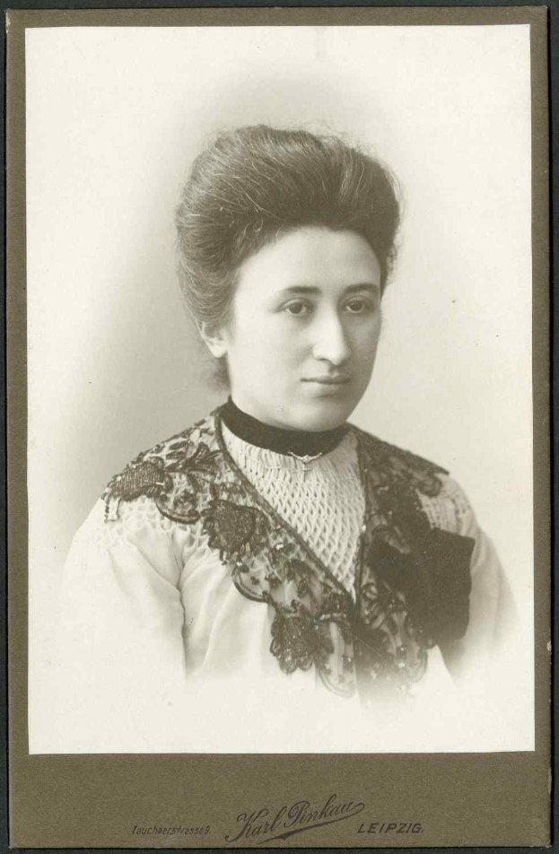 Schwarä-weißes Porträt einer Frau in einem weißen Kleid mit einem gemusterten Schultertuch, identifiziert als Karl Pinkau Leipzig, die direkt in die Kamera schaut.