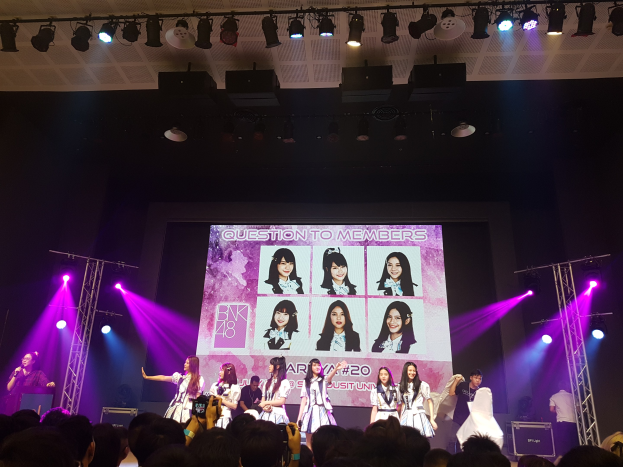 Eine Gruppe von Menschen tritt auf einer hell erleuchteten Bühne mit Trägern auf, während ein Sänger ein Mikrofon hölt und ein anderer ein Instrument spielt, während die Menge darunter das Konzert aufzeichnet; ein großer Bildschirm im Hintergrund zeigt Bilder der K-Pop-Gruppe BK48.