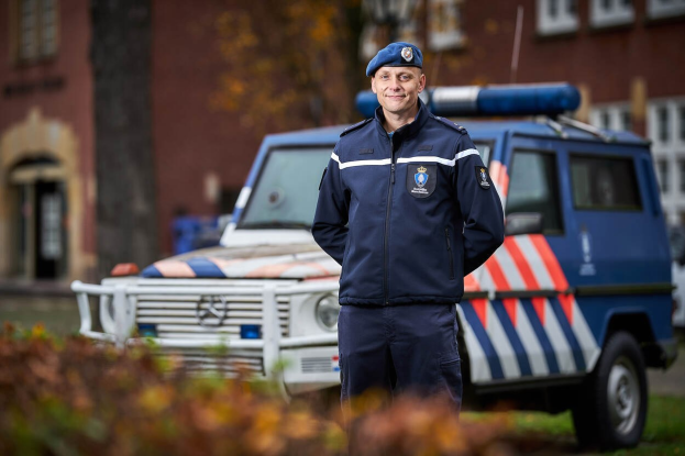 Ein Polizist in Uniform und Mütze steht vor einem Polizeiwagen mit leicht unscharfem Hintergrund und Pflanzen unten.