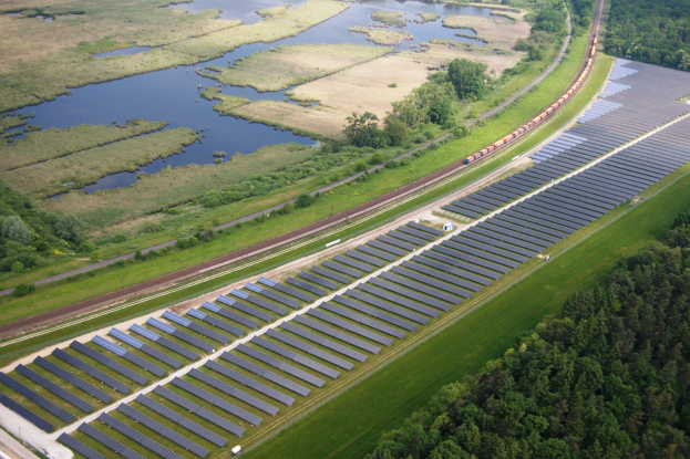 Luftaufnahme einer Solarpark mit Panelen, umgeben von Vegetation und Wasser, mit einem Zug auf einer nahen Schiene.