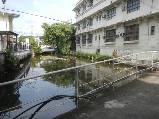 Ein kleiner Kanal verläuft durch eine Stadtstraße neben hohen Gebäuden, mit Wasser, Geländern, Rohren, Pflanzen, Bäumen, Strommasten mit Drähten und einem bewölkten Himmel im Hintergrund.