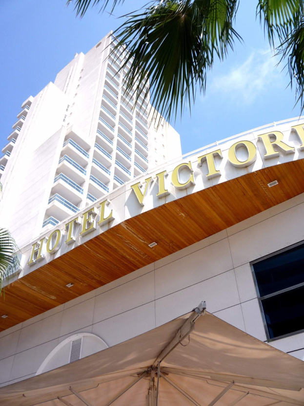 Das Bild zeigt das Hotel Victoria in Miami Beach, Florida, mit einem hohen Gebäude mit Glasfenstern und einem Namen auf der Wand, mit einem Zelt und einem Baum unten und einem Himmel voller weißer Wolken im Hintergrund.