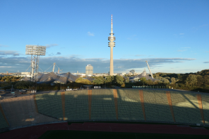 Luftaufnahme des Olympiastadions in Berlin, Deutschland, mit dem Fernsehturm (Fernsehturm) im Hintergrund, umgeben von Bäumen, Gebäuden und beleuchteten Bereichen unter einem bewölkten Himmel.