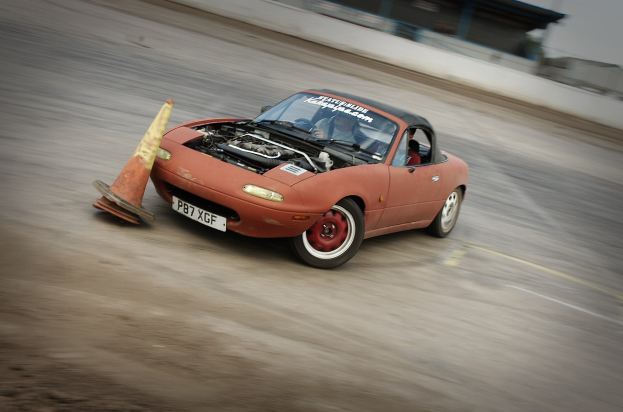 Roter Wagen auf einer Rennstrecke mit einem Verkehrskegel vorne, Hintergrund unscharf durch hohe Geschwindigkeit.