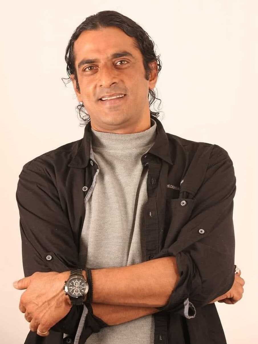 Ein Mann in einem schwarzen und aschgrauen Outfit mit verschränkten Armen, der eine Uhr trägt, steht vor einem weißen Hintergrund und sieht nachdenklich aus.