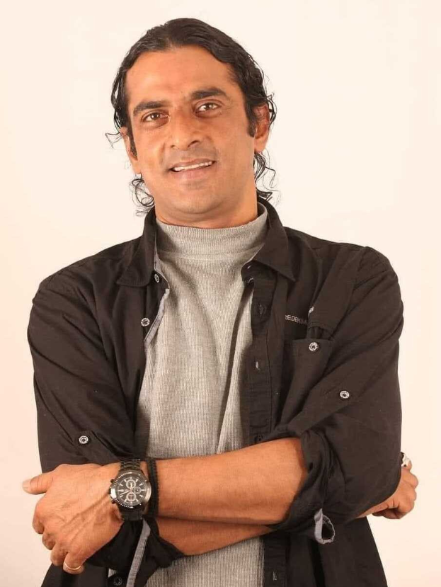 Ein Mann in einem schwarzen und aschgrauen Outfit mit verschränkten Armen, der eine Uhr trägt, steht vor einem weißen Hintergrund und sieht nachdenklich aus.