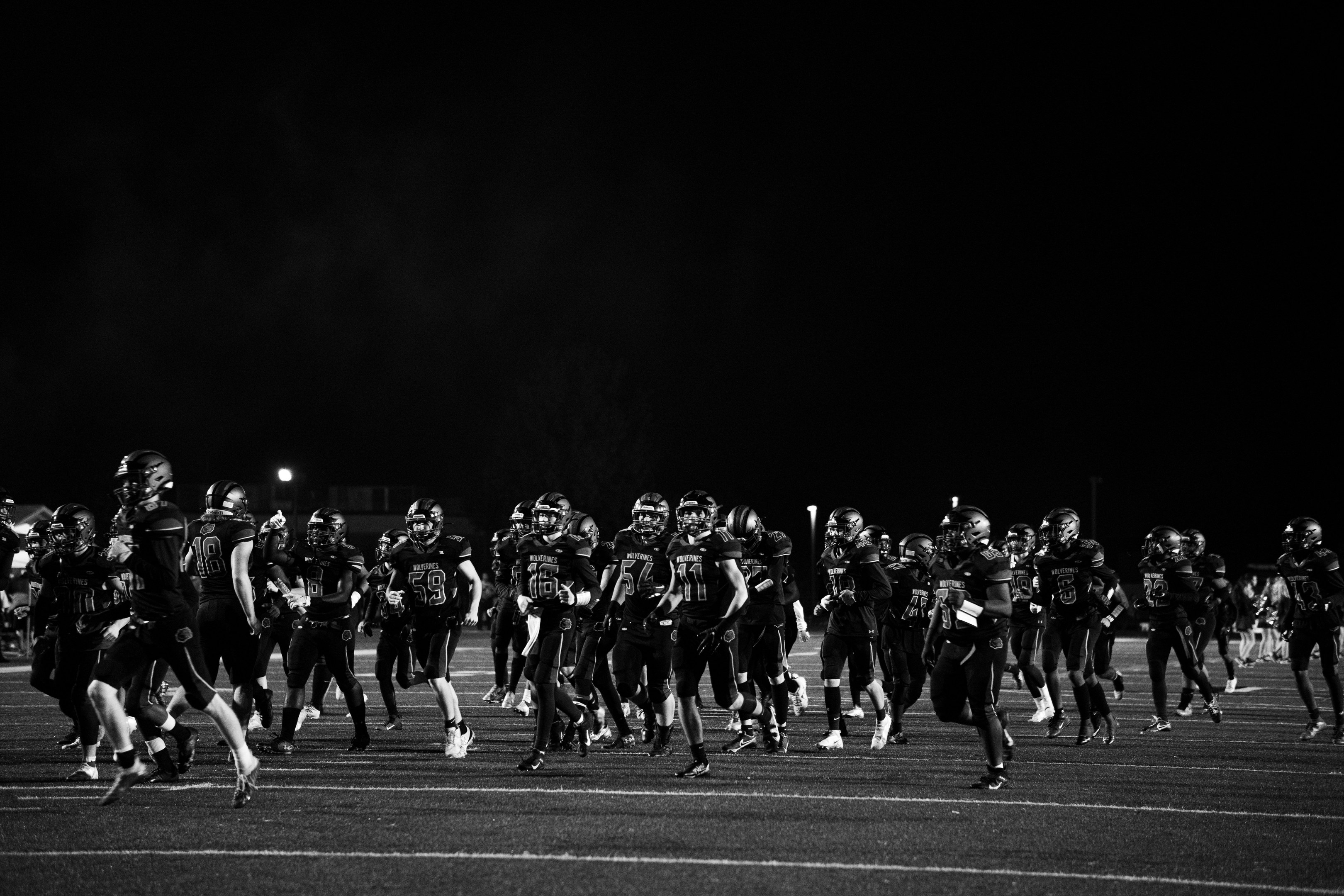 Schwarzes und weißes Bild von Fußballspielern in Helmen, die nachts auf das Feld laufen, umgeben von Stadionlichtern und Pfählen.