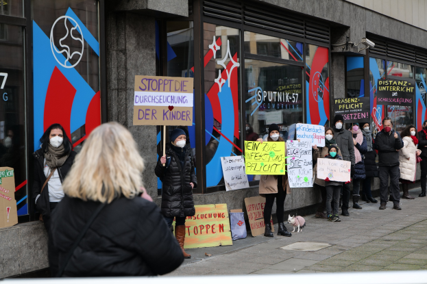 Maskierte Menschen mit Schildern und Plakaten protestieren vor einem Gebäude mit Glastüren, mit Kameras und einem Hund vor Ort.